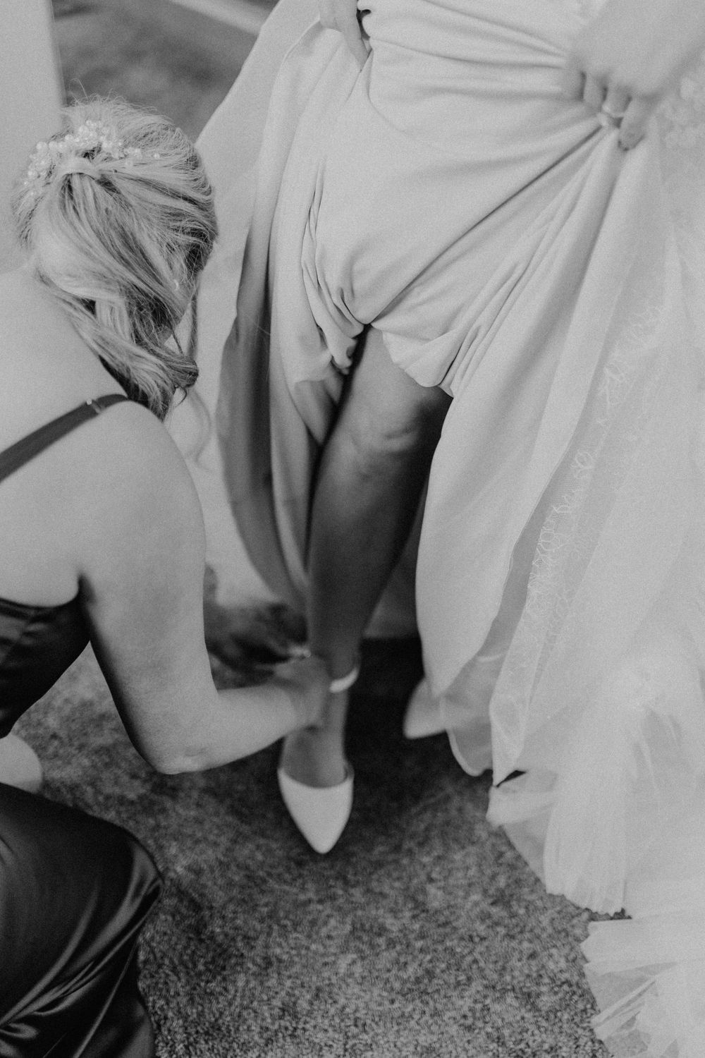 Bridesmaid helping bride put on white heels in black and white wedding preparation photo.