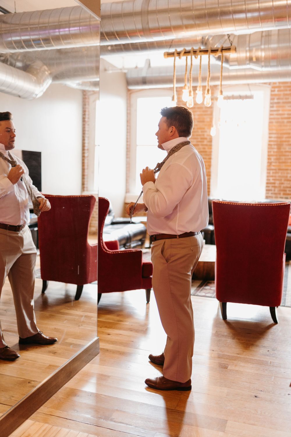 Groom adjusting his tie while getting ready, reflected in a mirror in a modern loft space.