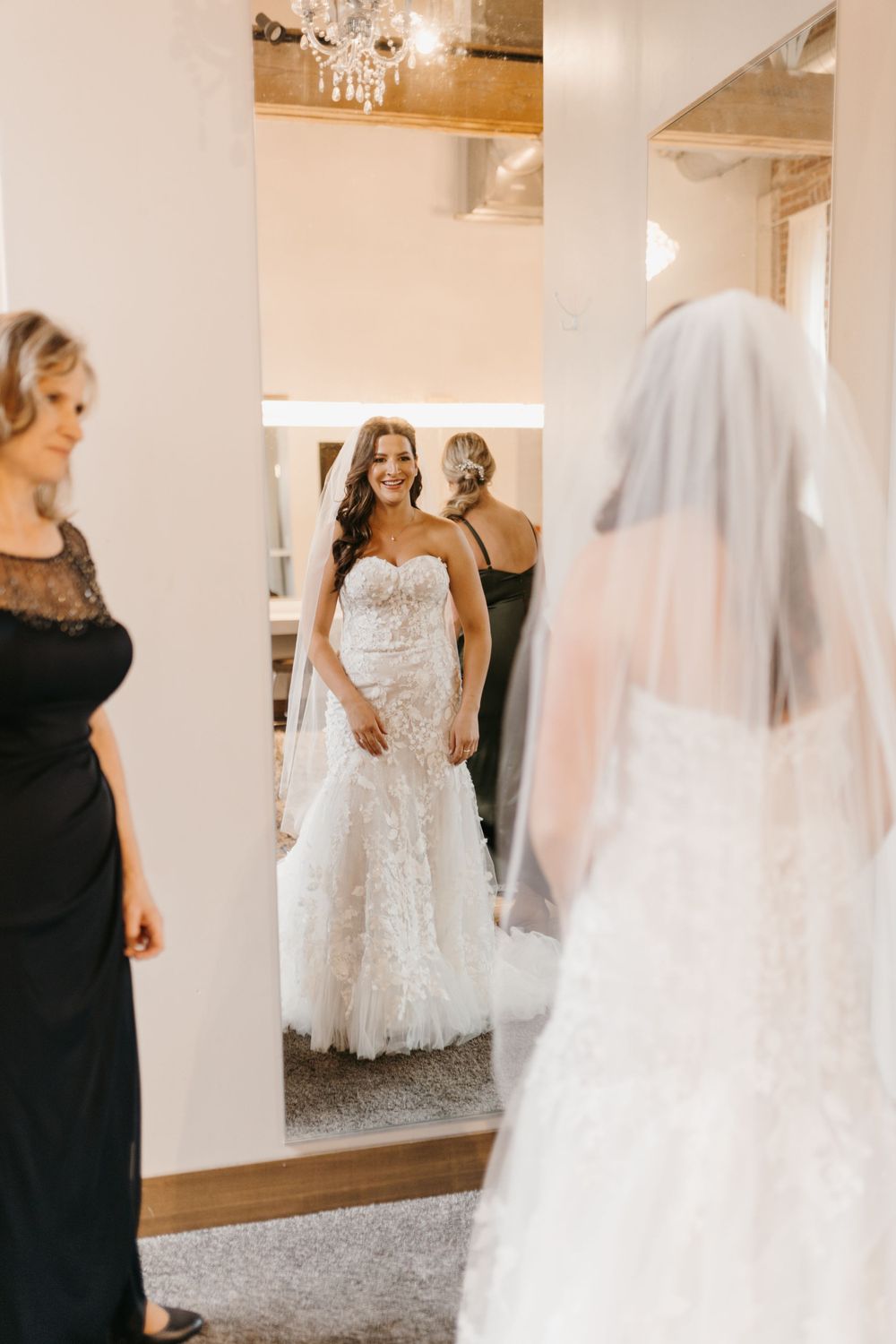 Bride in strapless lace mermaid wedding gown smiling at her reflection in a mirror surrounded by bridesmaids.
