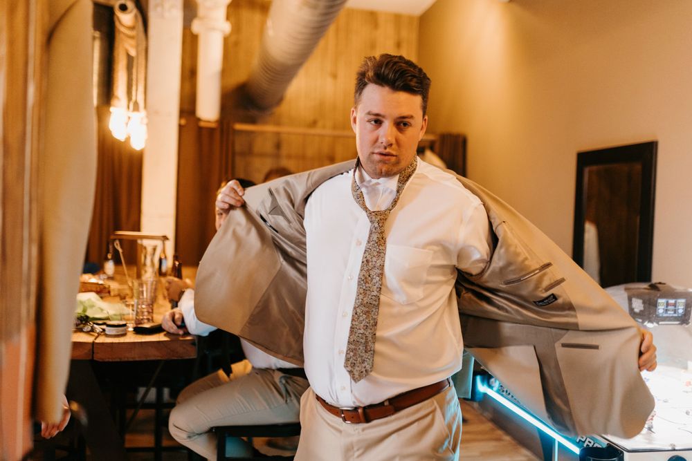 Groom putting on his tan suit jacket while getting ready for his wedding day.
