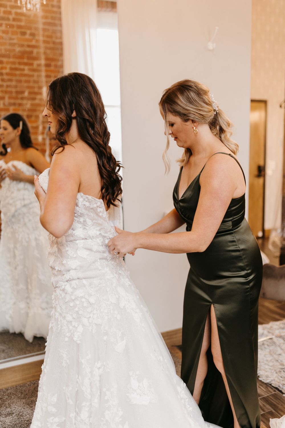 Bridesmaid in olive green satin dress helping bride button up her lace wedding gown.