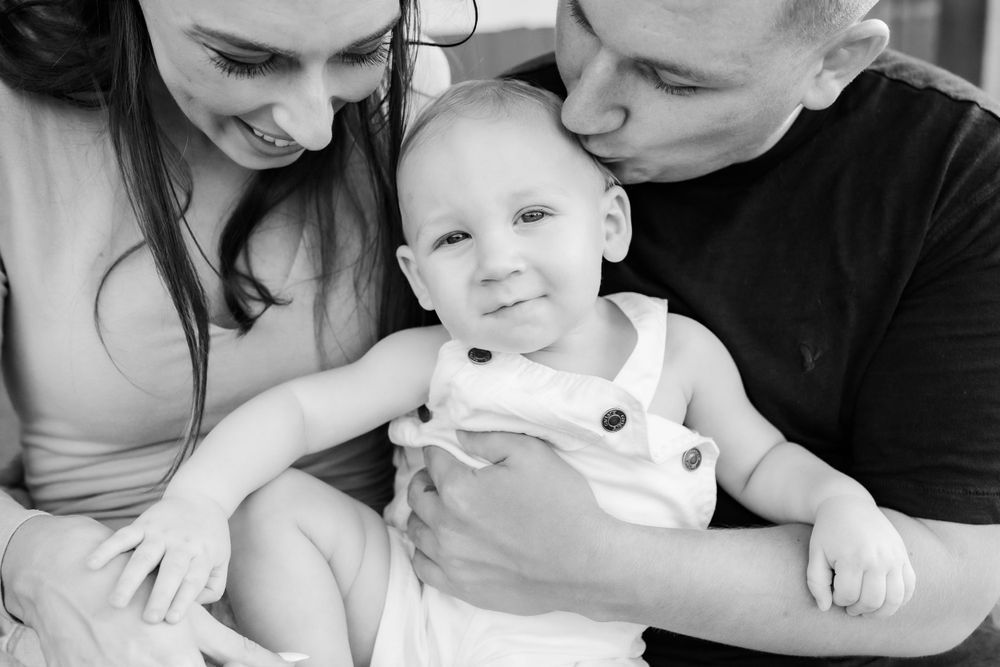 Black and white photo of loving parents kissing and cuddling their smiling baby.