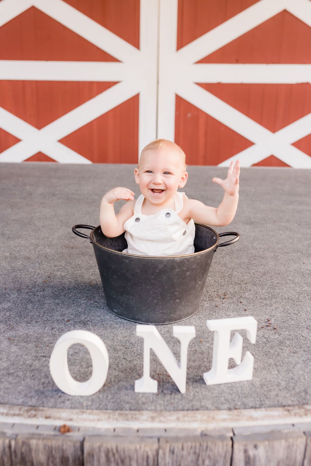 Laughing baby in metal tub with ONE letters in front of red barn for first birthday.