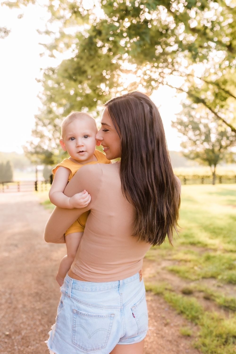 Mother kissing baby on cheek while holding infant outdoors in golden sunset light.