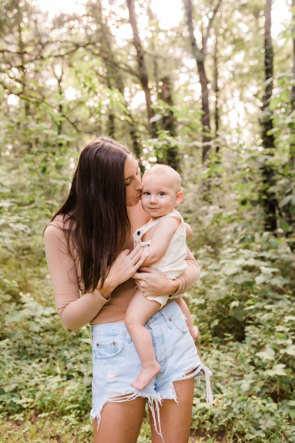Mother with long dark hair kissing her smiling baby while standing in a sunlit green forest.
