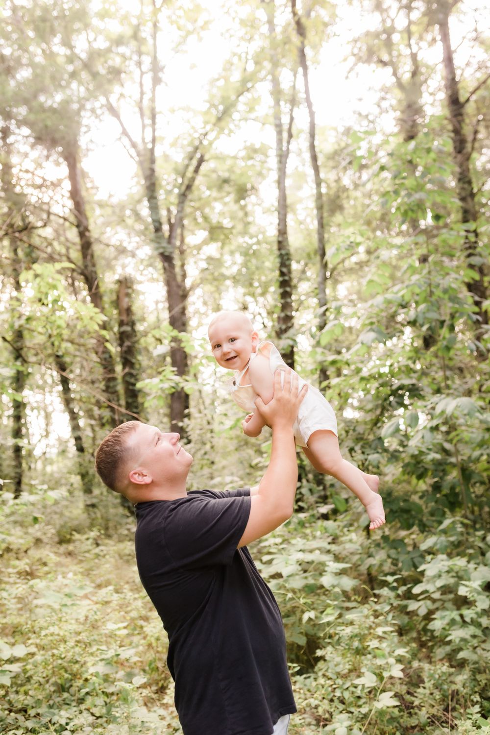 Joyful father lifting laughing baby high in the air surrounded by lush green trees in sunlight.