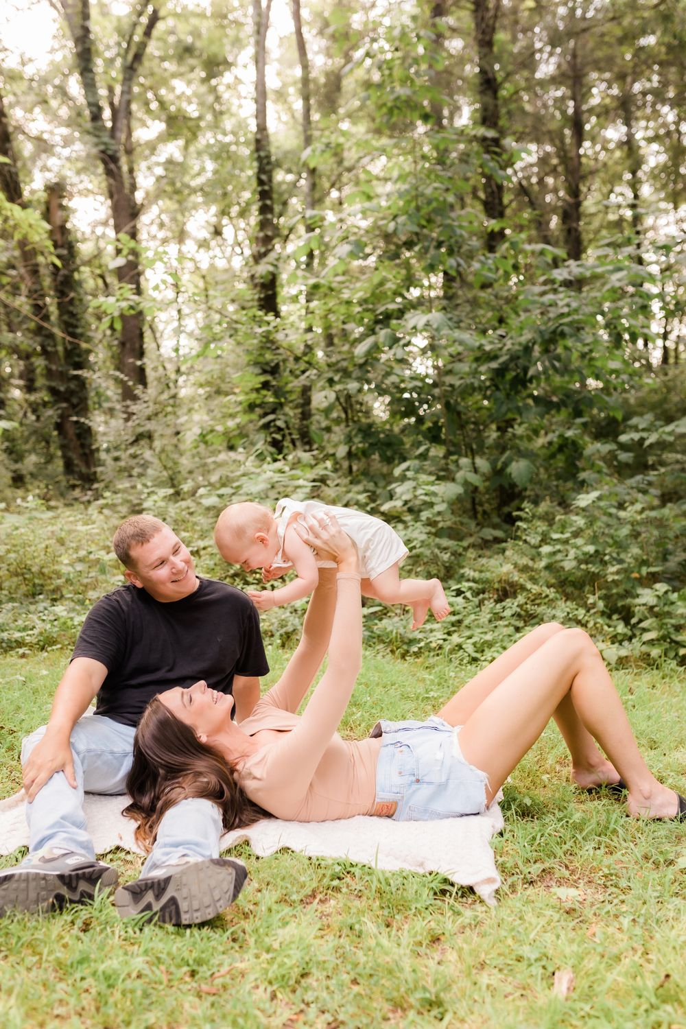 Happy family lying on a blanket in the grass as mother lifts smiling baby in the air outdoors.