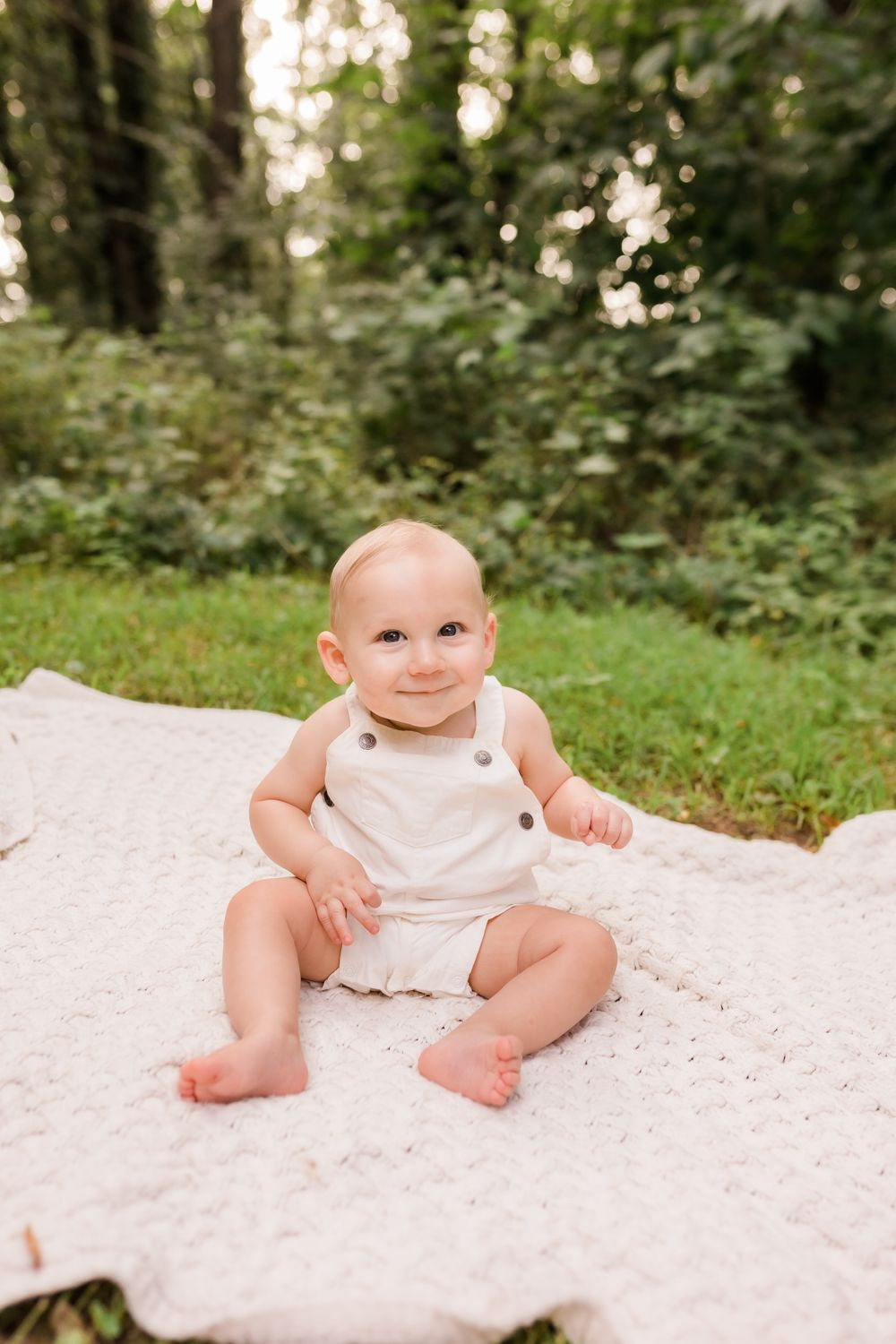 Smiling baby sitting on white blanket outdoors in lush green garden wearing white romper.