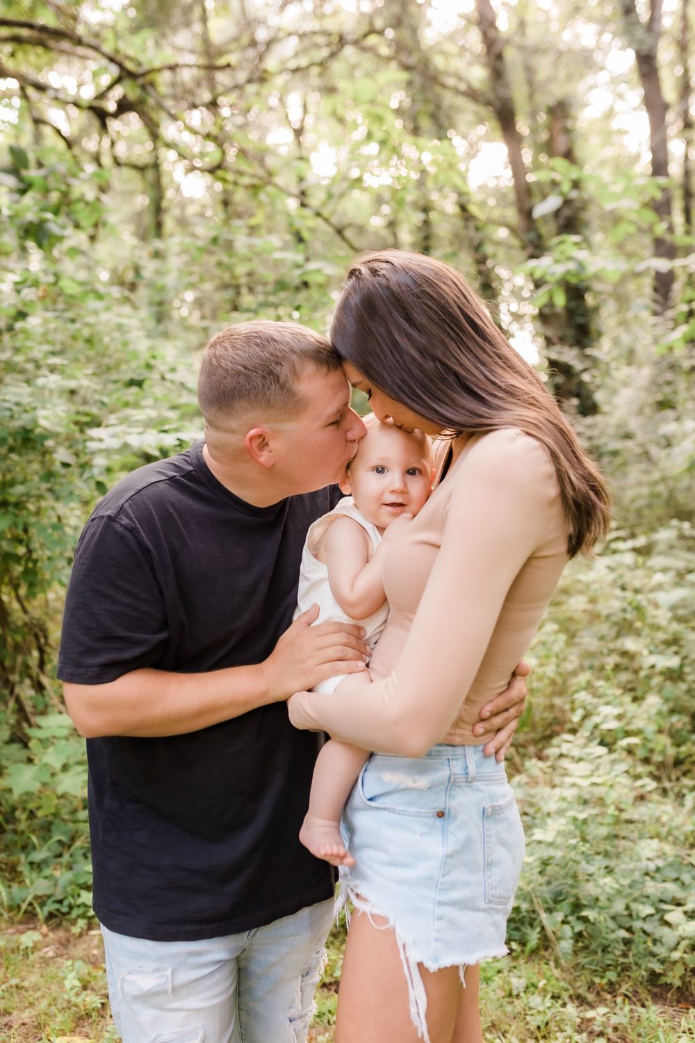 Young family of three with parents kissing their baby on the cheeks in a lush green forest setting.