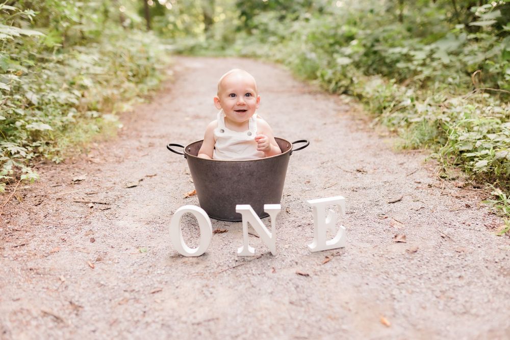 Happy baby sitting in metal tub on dirt path with ONE letters for first birthday photos.