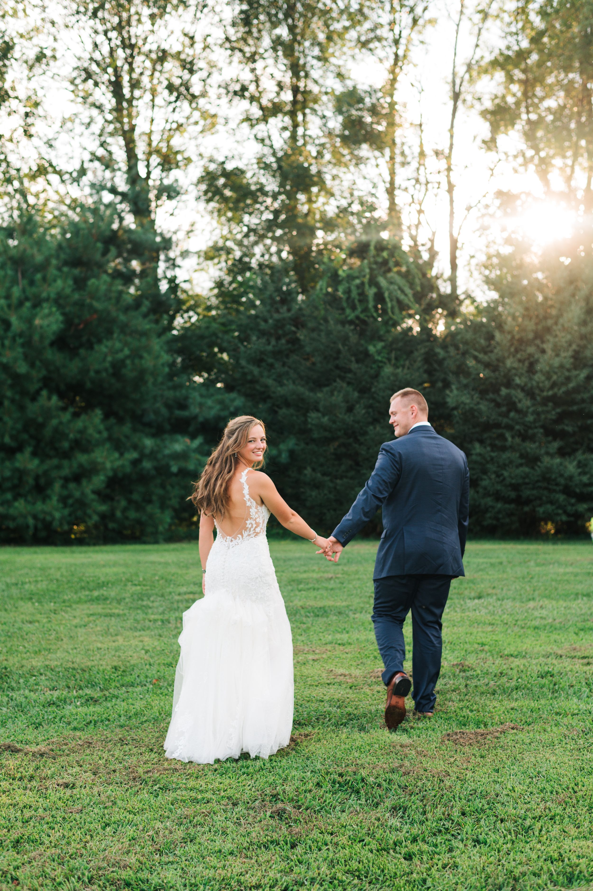 A romantic wedding couple walks hand in hand across a sunlit lawn surrounded by tall trees.