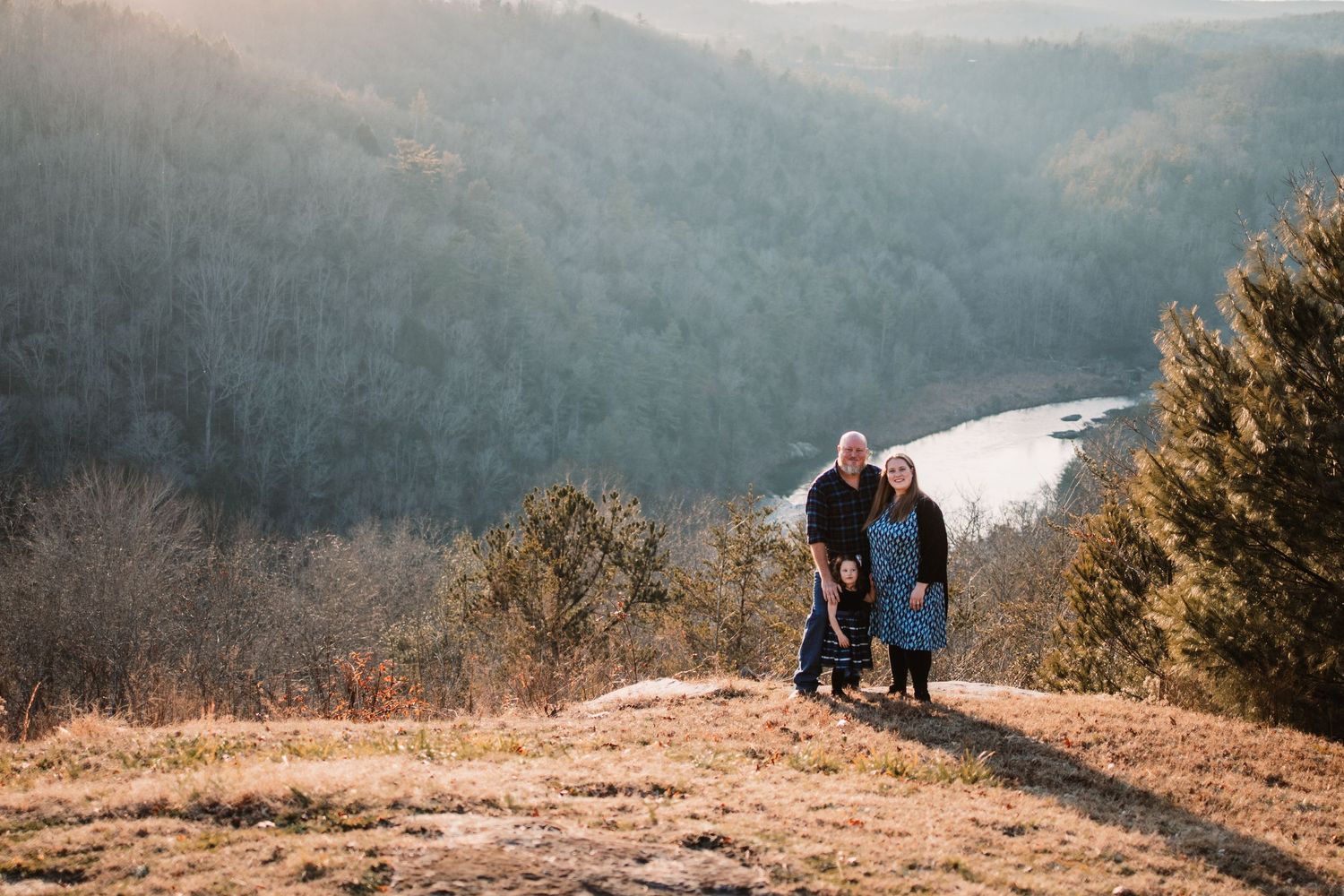 Couple standing on a rocky overlook surrounded by forested hills and a river valley at sunset.