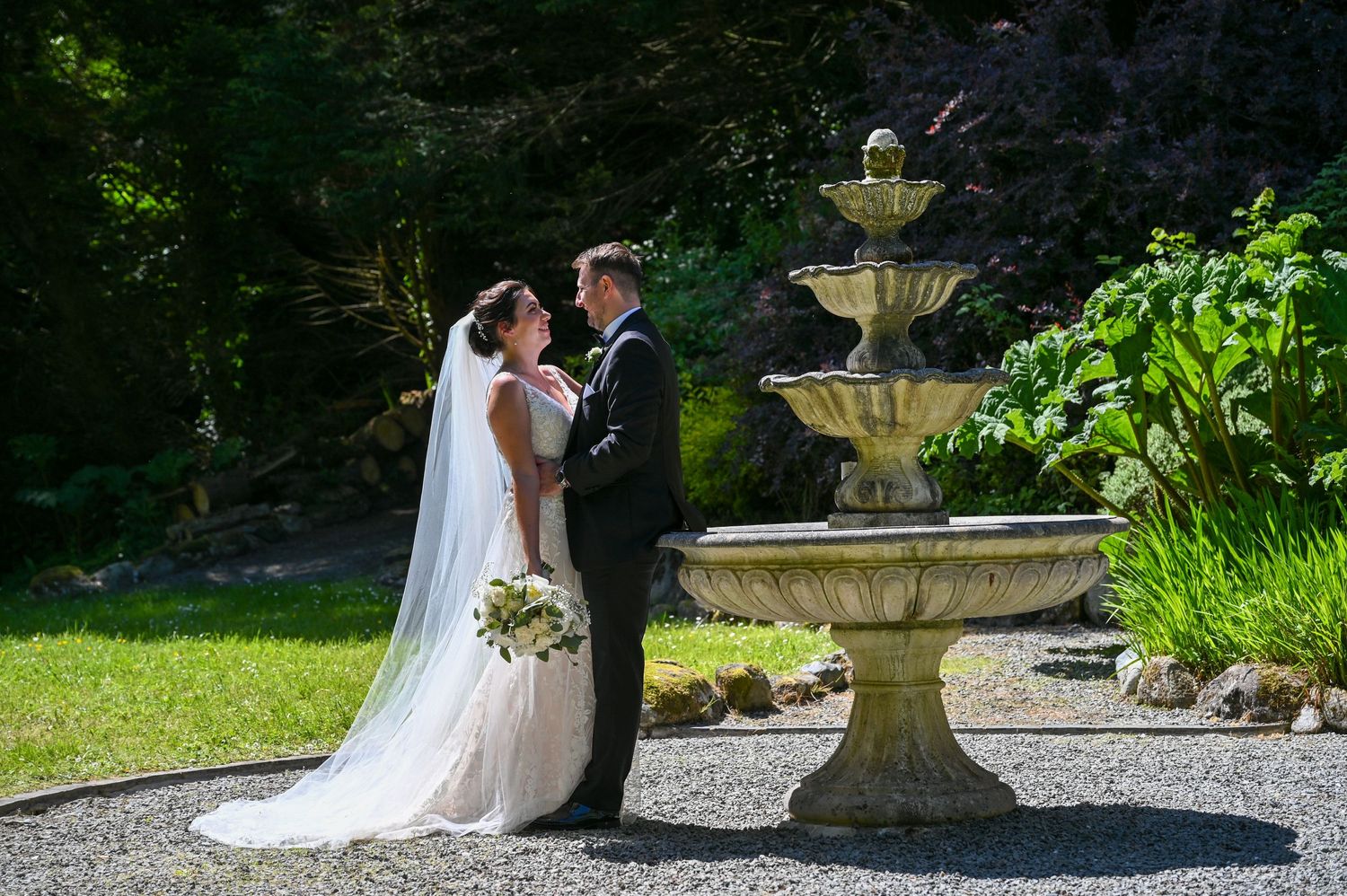 Newlyweds pose together by an ornate garden fountain with the bride holding a white bouquet.