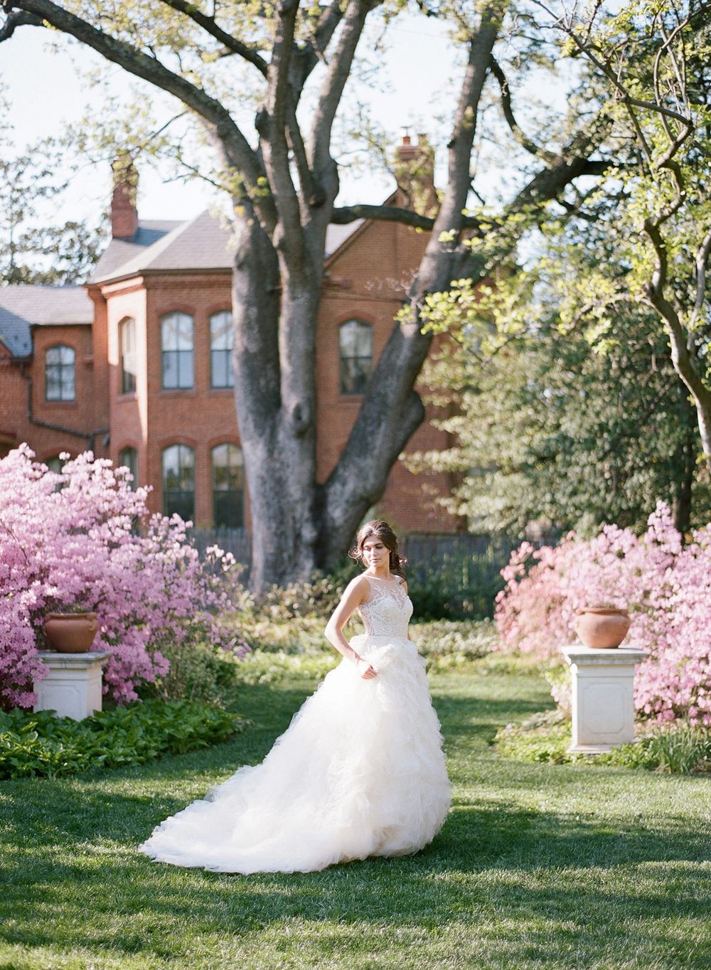 A bridal portrait in a garden with blooming pink cherry blossoms and a grand brick mansion in the background.