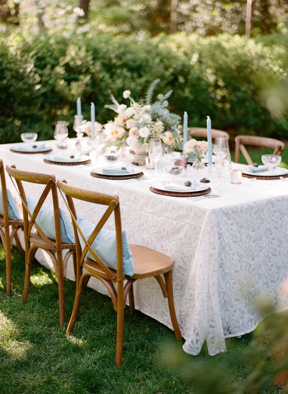 Elegant outdoor table setting with white lace tablecloth, wooden cross-back chairs, and delicate floral centerpieces.