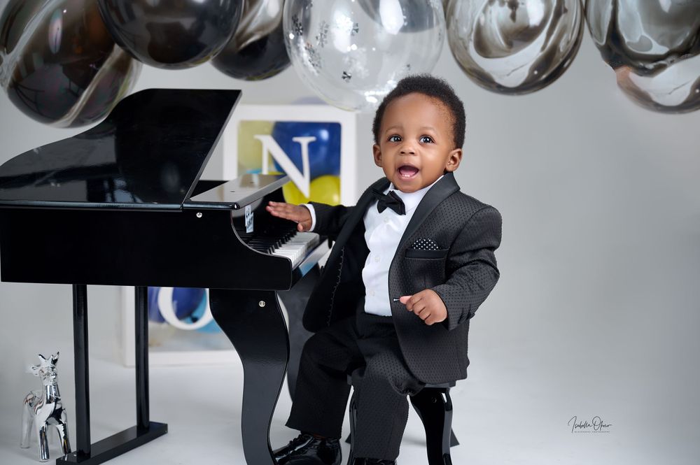Young child in formal black tuxedo and bowtie poses playfully next to a grand piano during professional photoshoot.