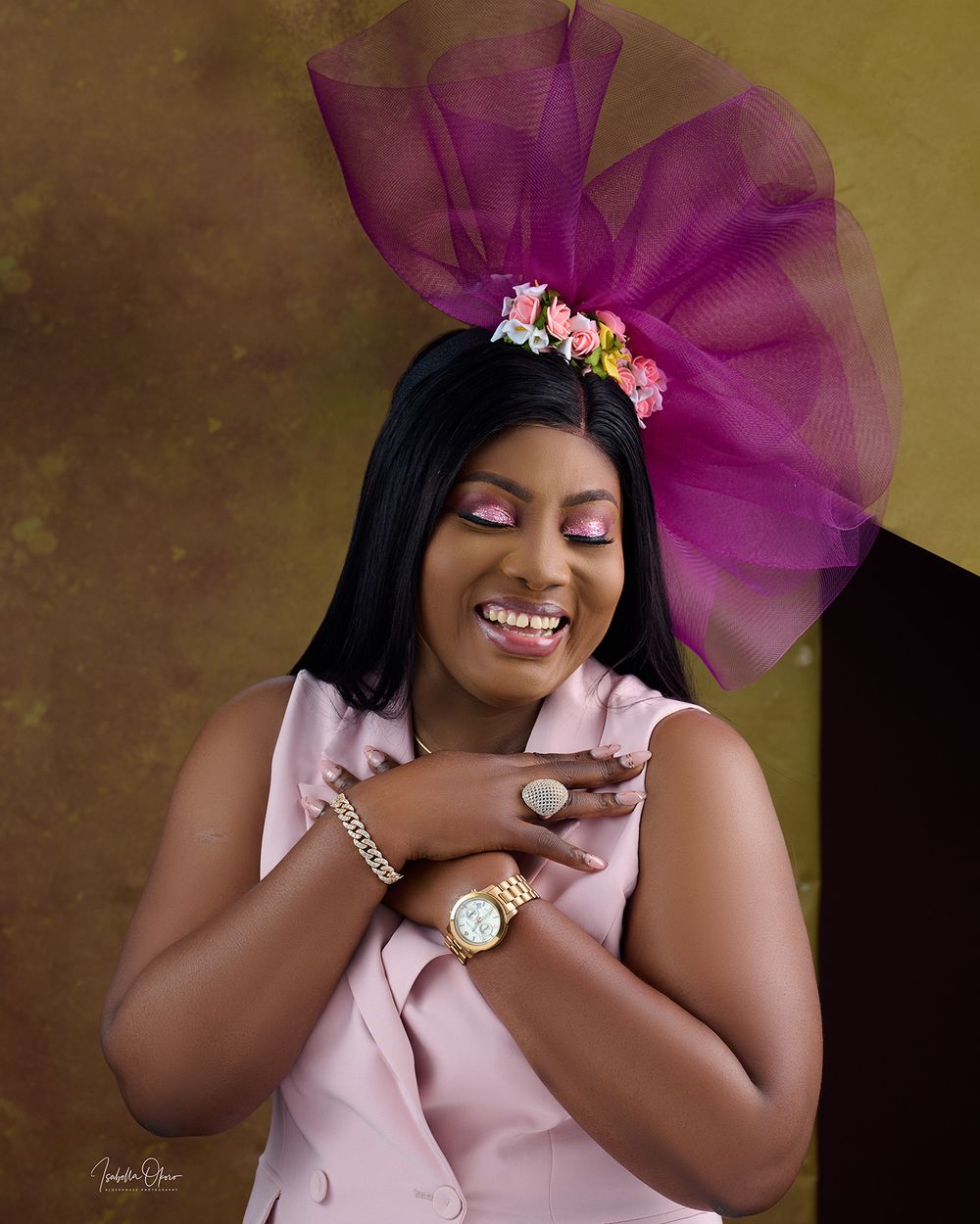 Professional portrait of a woman in pink wearing a purple floral headpiece and tulle against a gold backdrop.