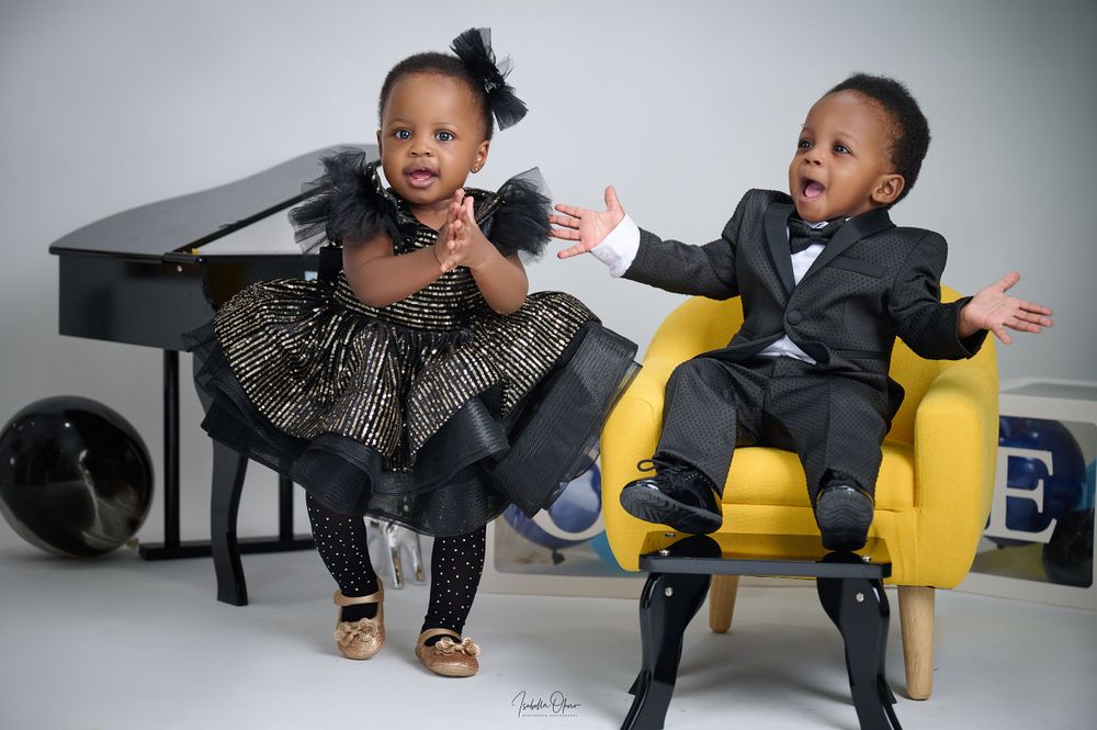 Two children in formal attire pose expressively by a piano in a studio setting.