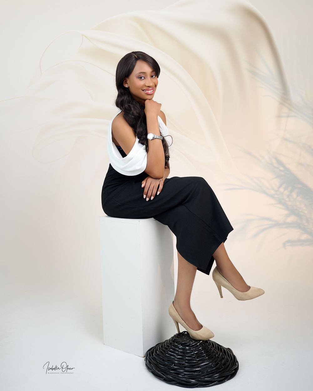 Stylish portrait of a woman in black and white outfit seated on pedestal against beige background.