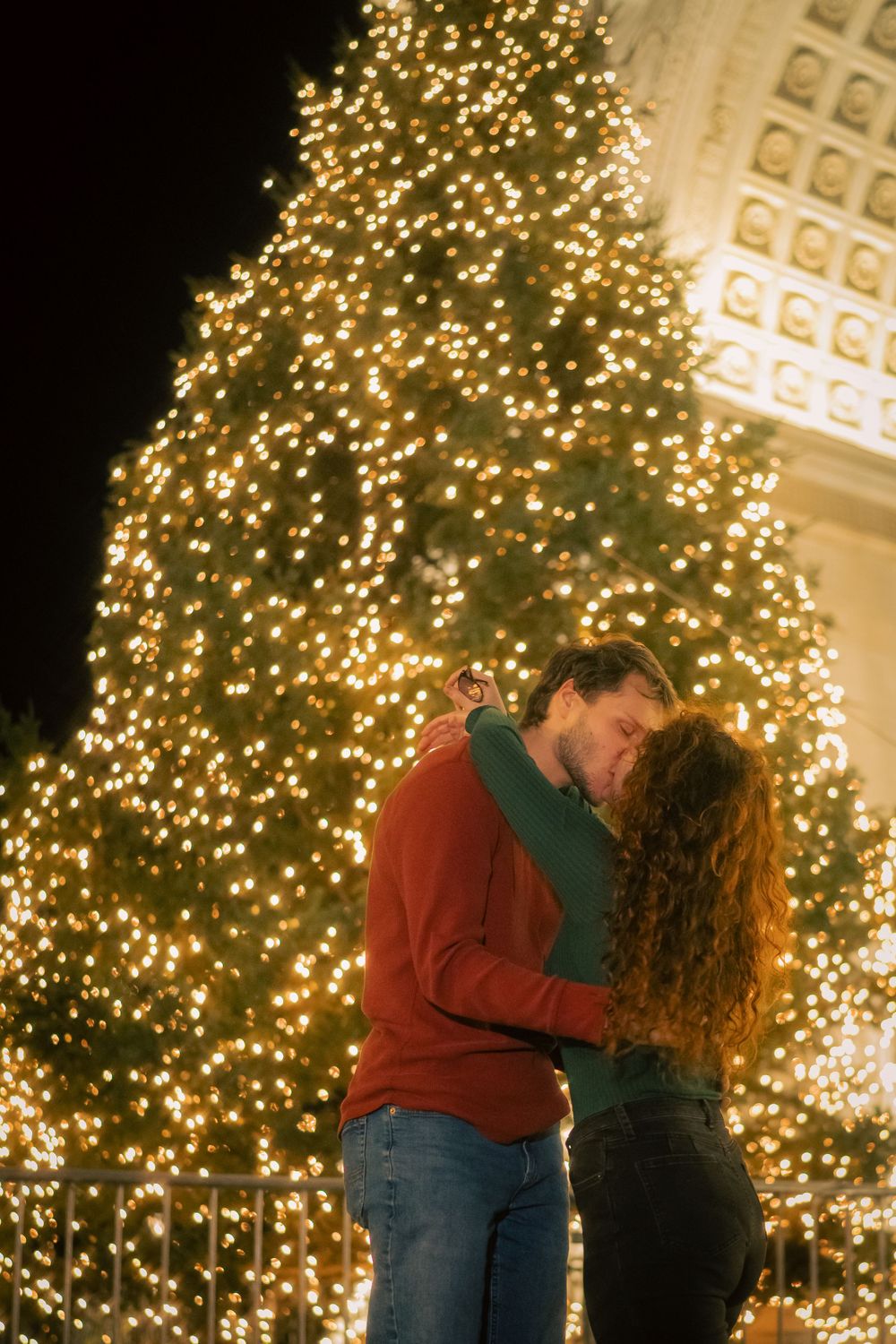 A couple embraces near a large illuminated Christmas tree during evening.