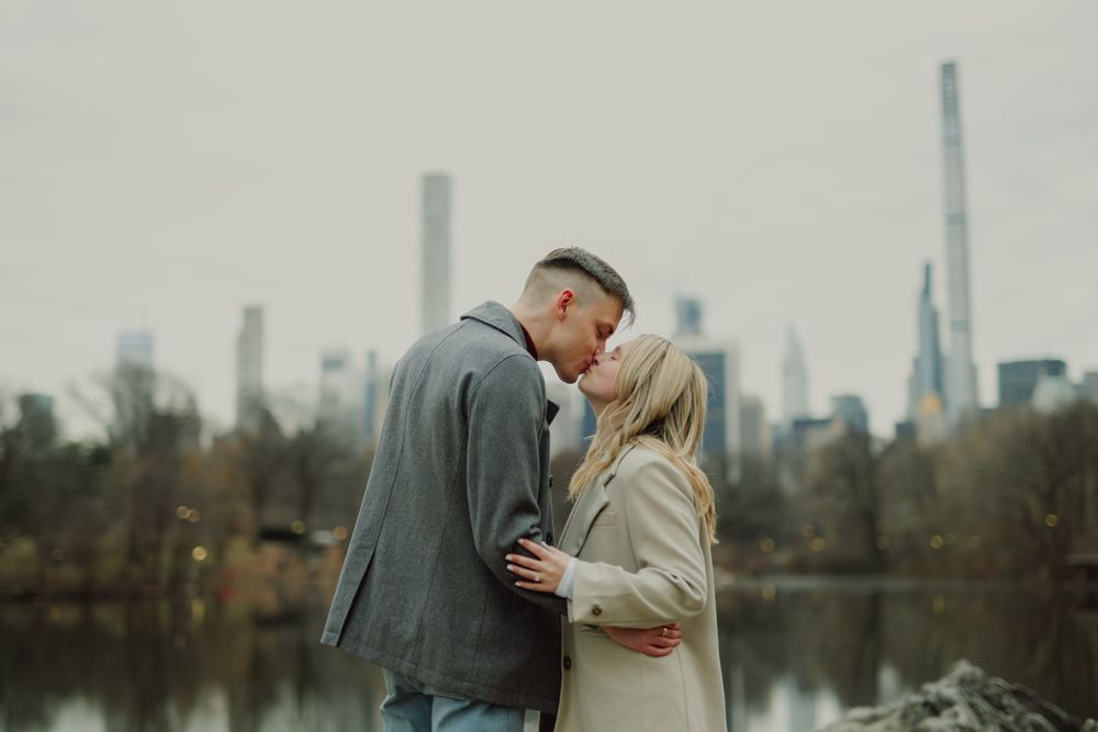 A couple embraces while standing by a lake with urban skyline visible in the distance.