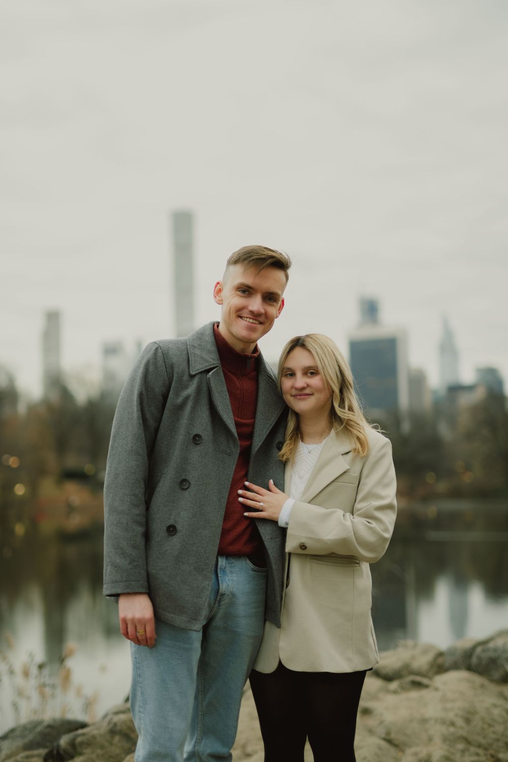 Two people embrace while standing on rocky terrain near a lake with city buildings visible.