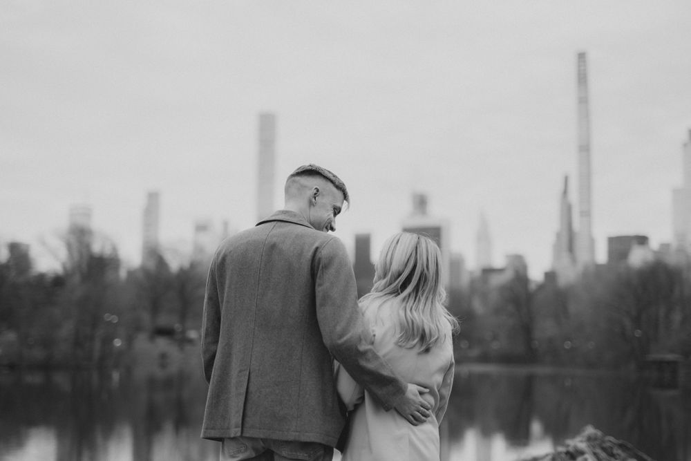 A couple embraces near a waterfront with urban skyline visible in the distance.