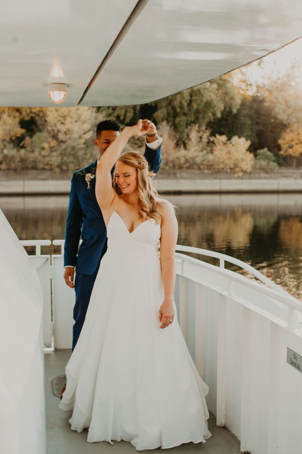 Groom twirls smiling bride in white wedding gown on a sun-drenched boat deck by the water.