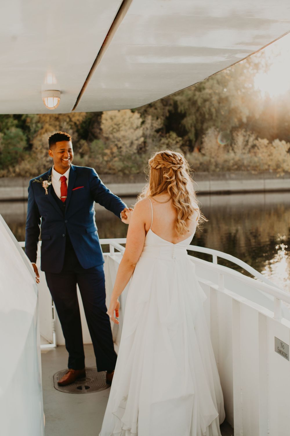 Groom in navy suit reaches for bride's hand on sunlit boat deck during wedding photo session.