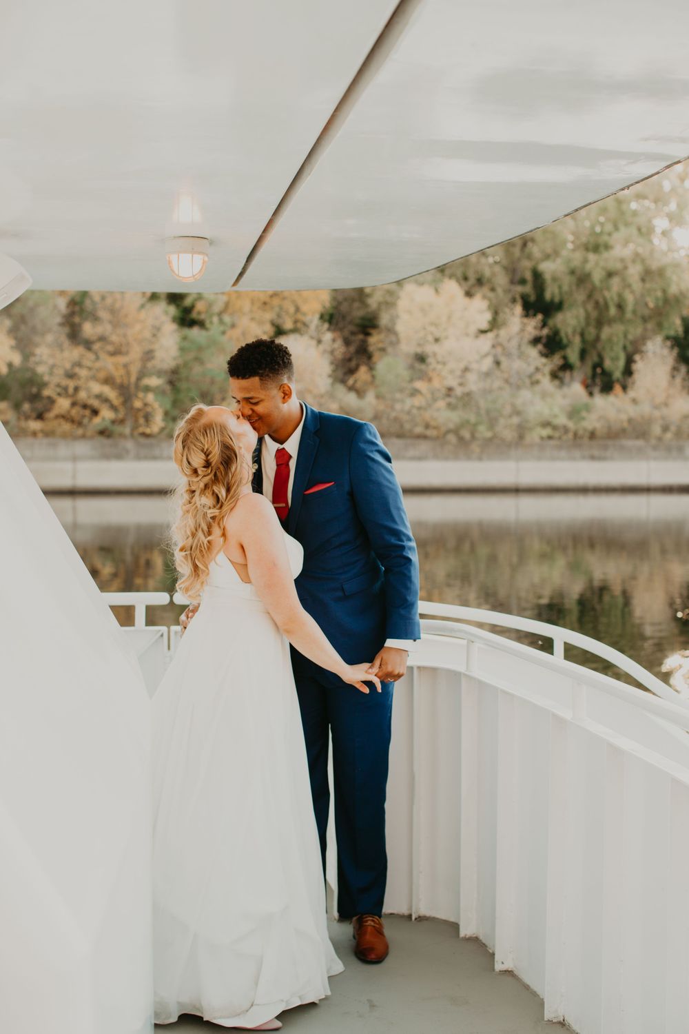 Bride and groom share a tender moment on a white boat deck surrounded by autumn foliage.