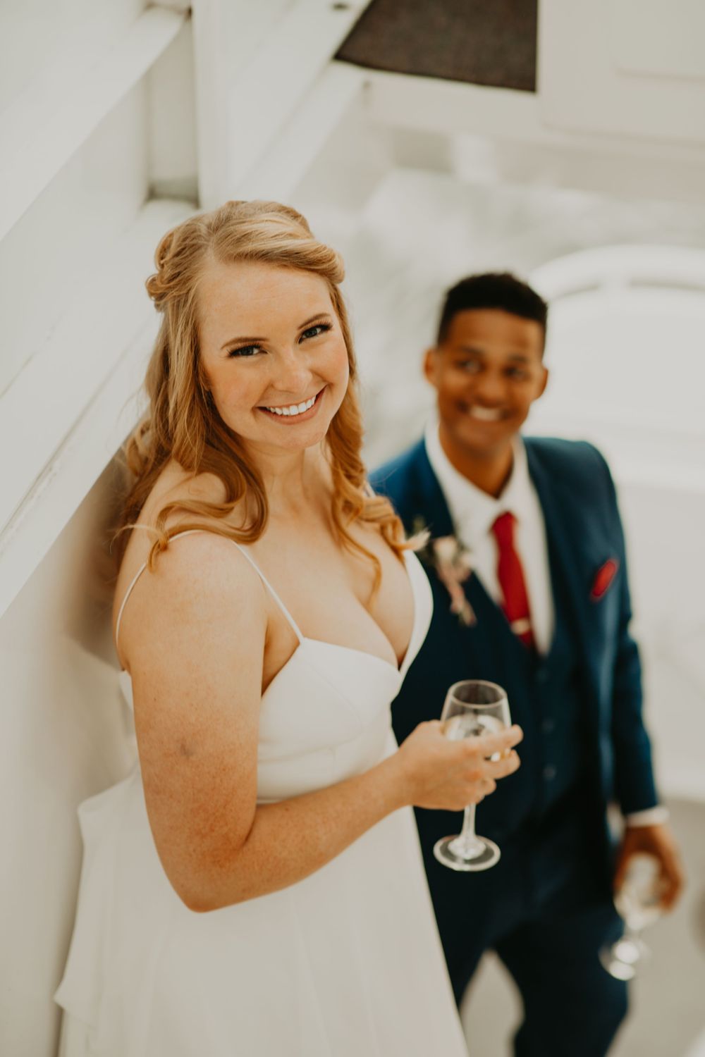 Radiant bride holds champagne glass and smiles on boat stairs with groom in navy suit behind her.