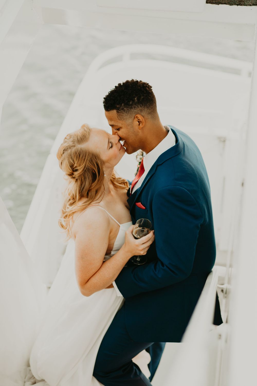 Newlyweds share a joyful kiss on a white boat, bride holding a champagne glass in golden light.