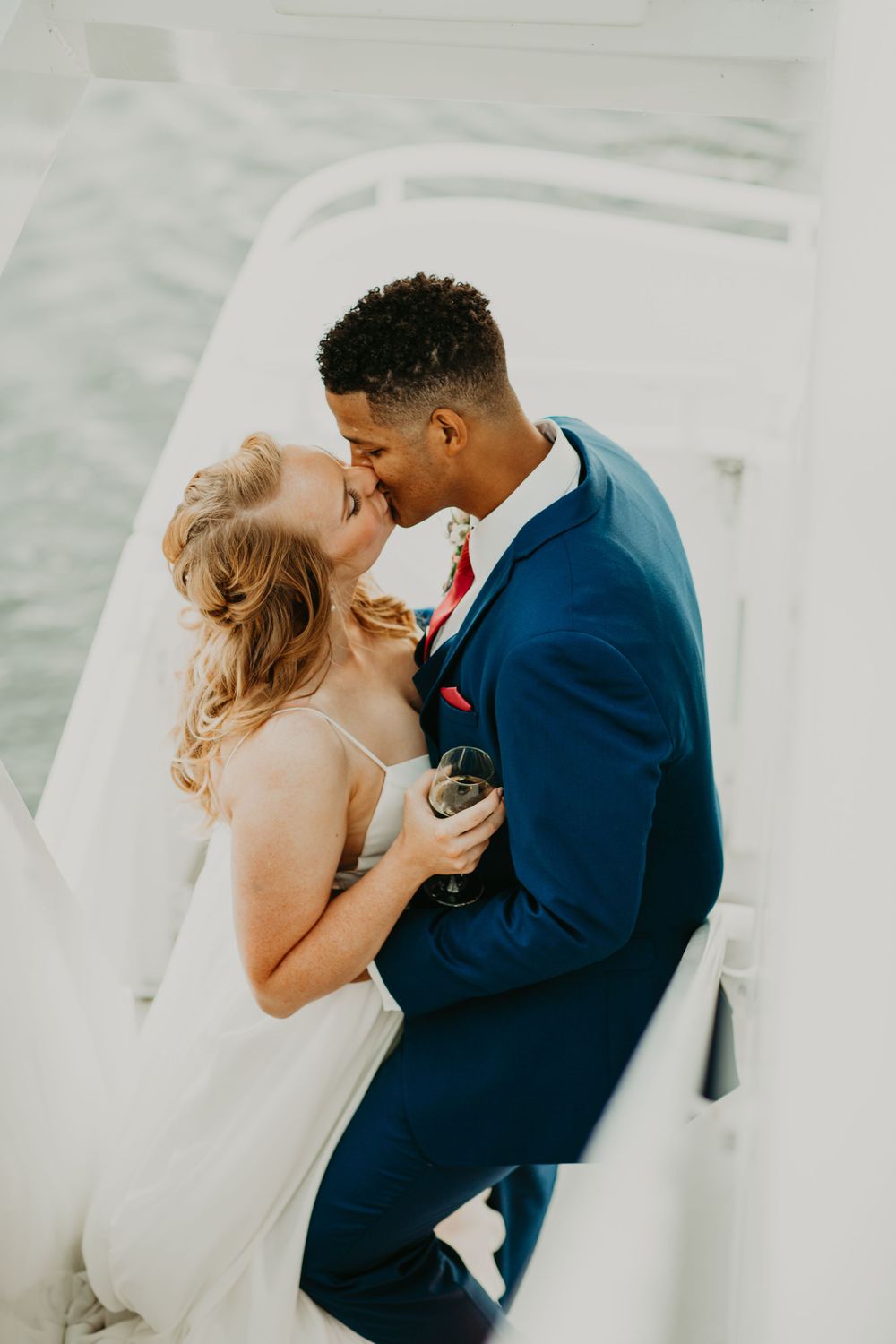 Interracial couple shares a romantic kiss on a white boat deck, groom in navy suit holding champagne.
