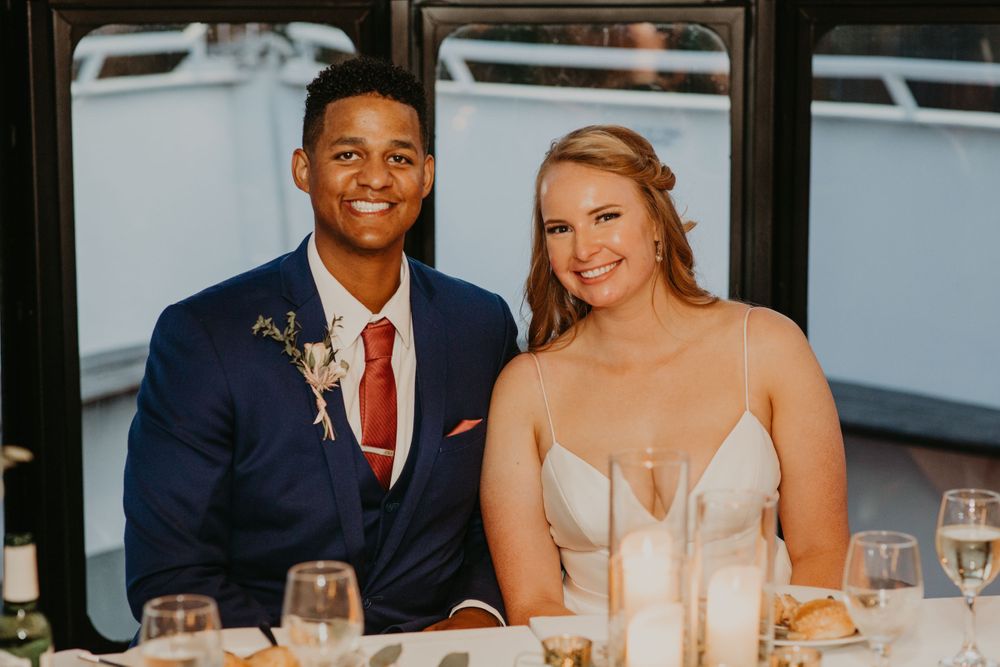 Bride and groom smiling at wedding reception table on a boat, groom in navy suit with red tie.