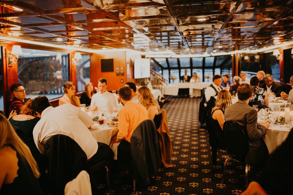 Elegant wedding reception dinner on a boat with guests seated at round tables under warm lights.