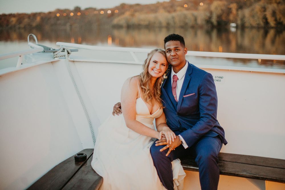 Wedding couple embracing on boat deck at golden hour with scenic waterfront background.