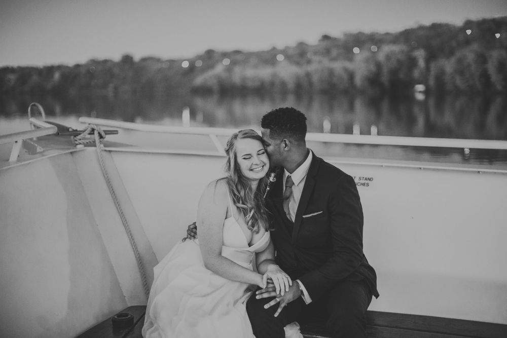 Black and white photo of smiling bride and groom sitting together on boat deck at dusk.