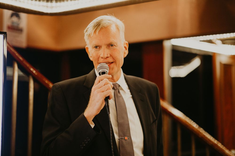 Older gentleman in dark suit giving a wedding speech into microphone at elegant venue.