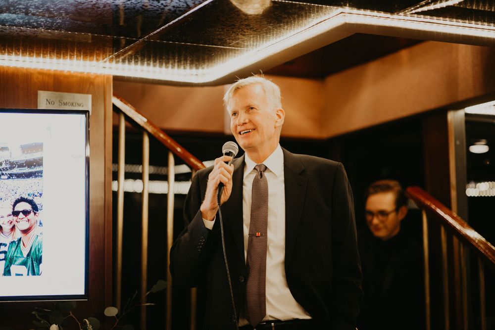 Man in dark suit delivering wedding toast with microphone near staircase at reception.