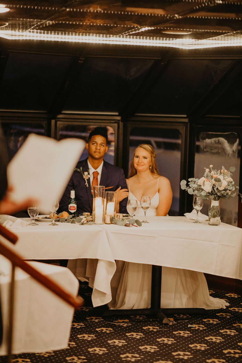 Interracial newlywed couple smiling at sweetheart table during wedding reception dinner.