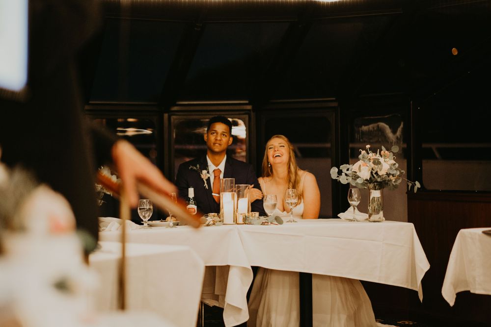 Joyful bride and groom laughing together at sweetheart table during wedding reception speeches.
