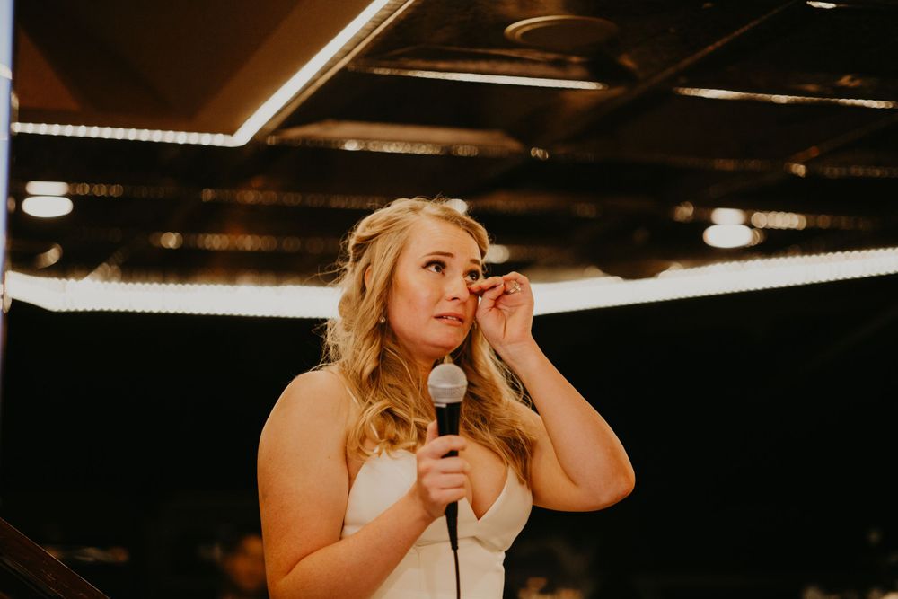 Emotional blonde bridesmaid wiping tear while giving heartfelt speech at wedding reception.