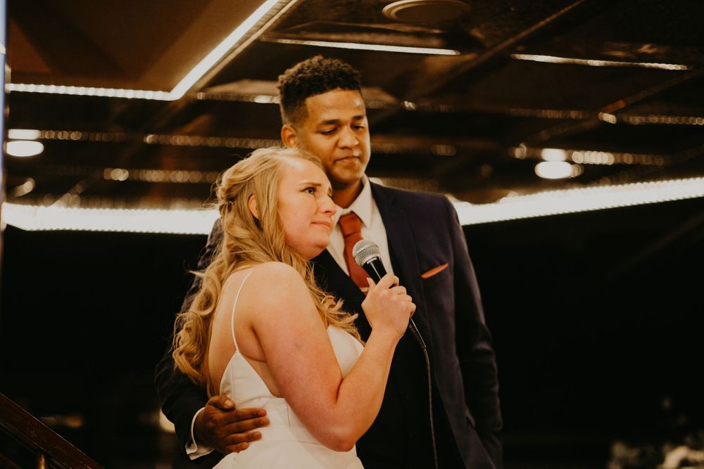 Groom in navy suit embracing bride from behind as she speaks into microphone at reception.