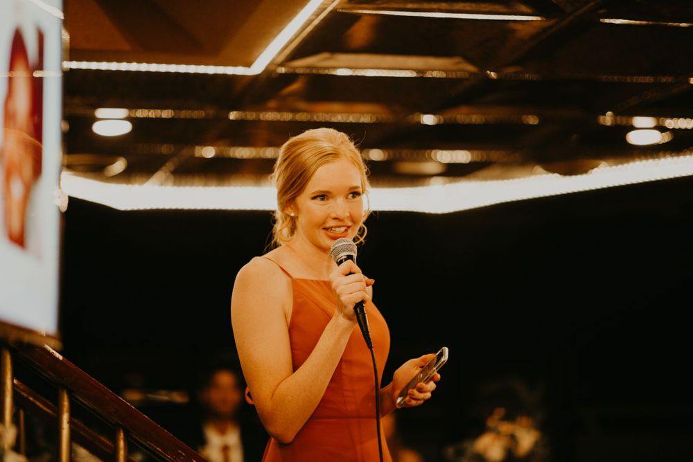 Elegant bridesmaid in rust orange dress giving wedding toast while holding microphone and phone.