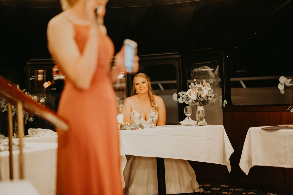 Smiling bride at sweetheart table watching bridesmaid in coral dress give wedding toast.