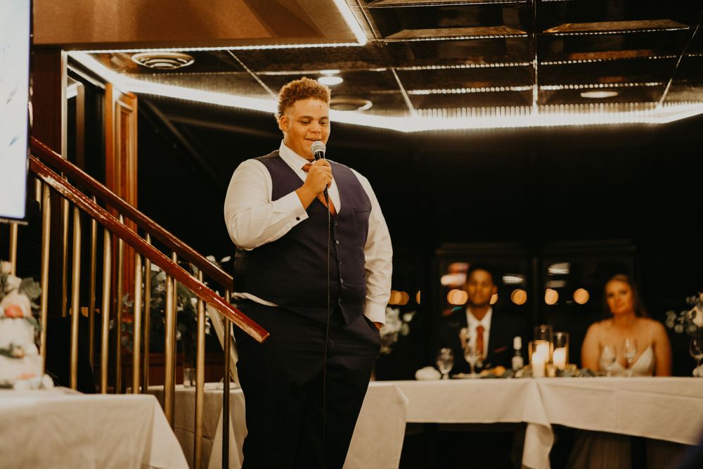 Groomsman in navy vest delivering heartfelt wedding speech near staircase at reception venue.