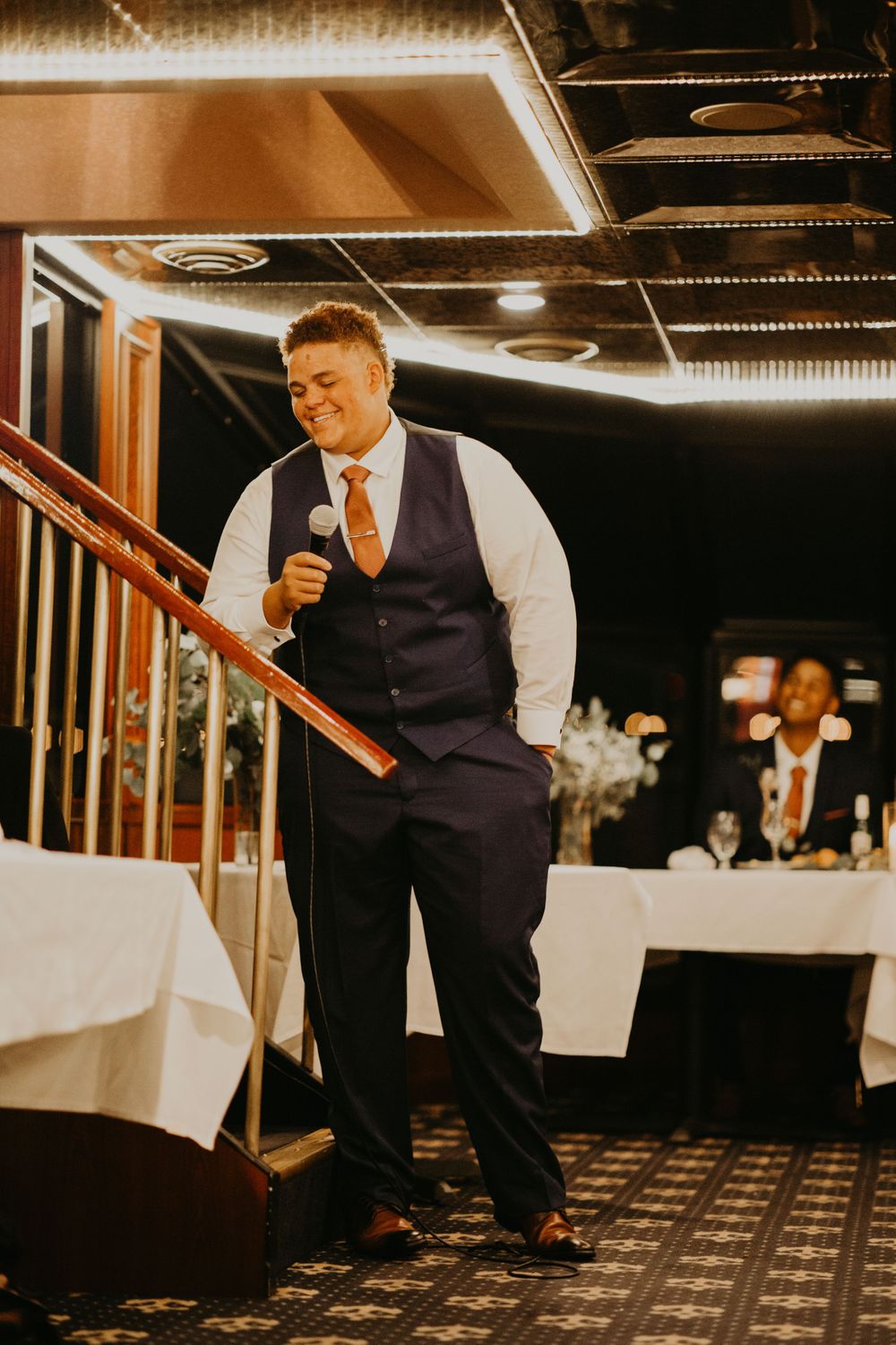 Groomsman in navy vest laughing while giving entertaining wedding speech near decorated staircase.
