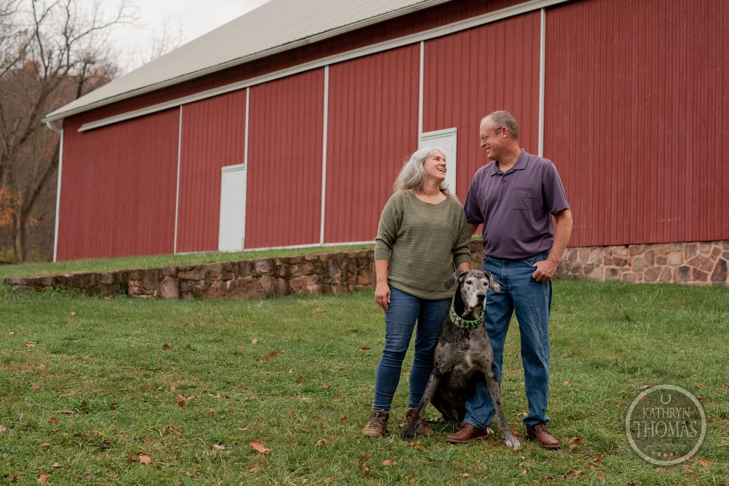 A couple and their black dog stand in front of a large red barn on a cloudy day.