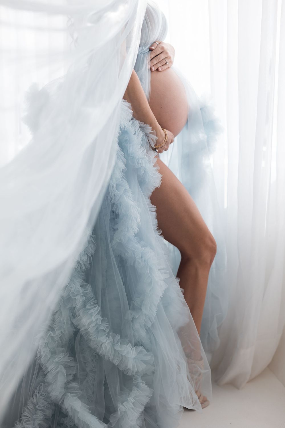 Pregnant woman posing behind sheer white curtains wearing flowing blue ruffled maternity gown.