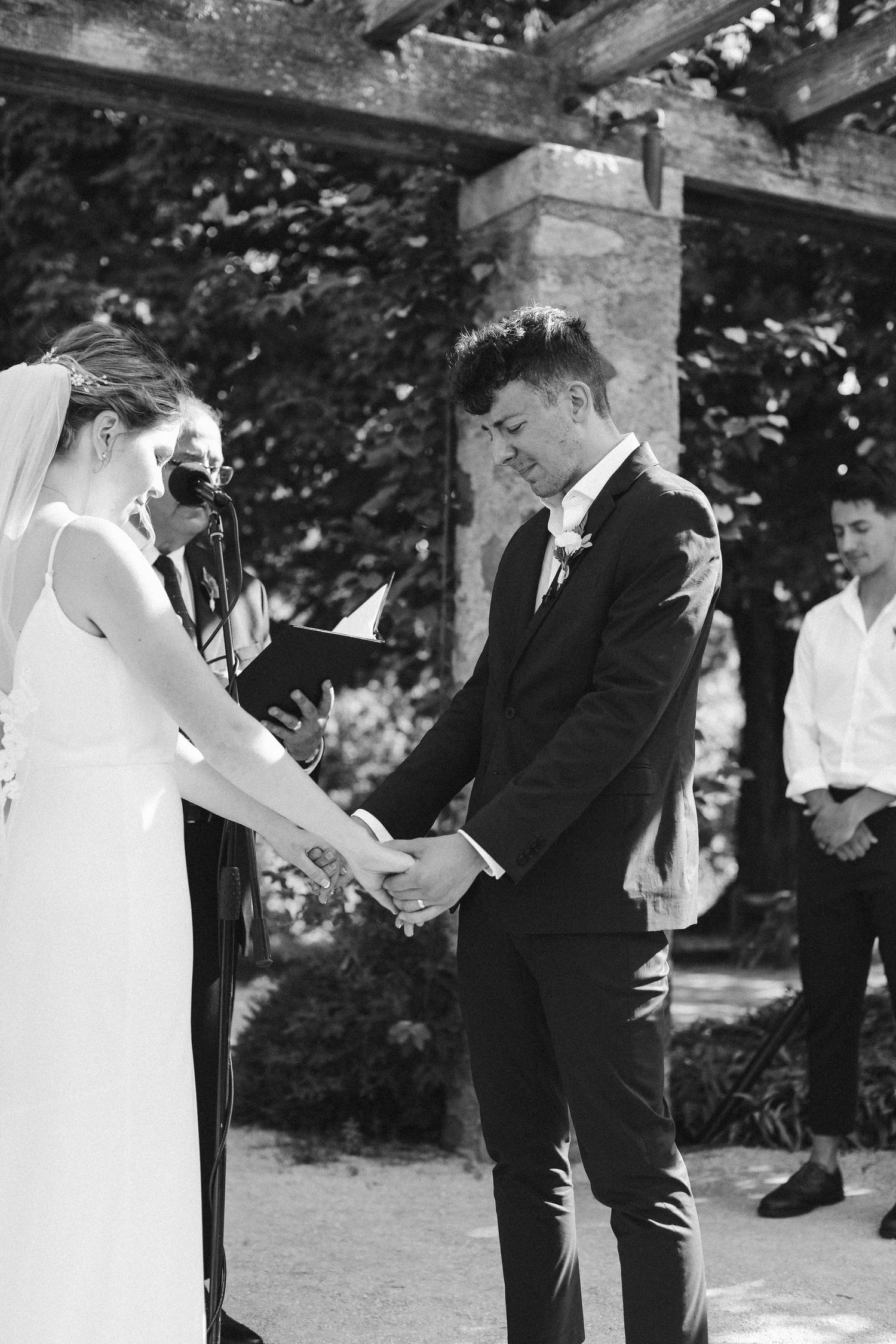 Black and white ceremony photo of couple holding hands during outdoor wedding vows.